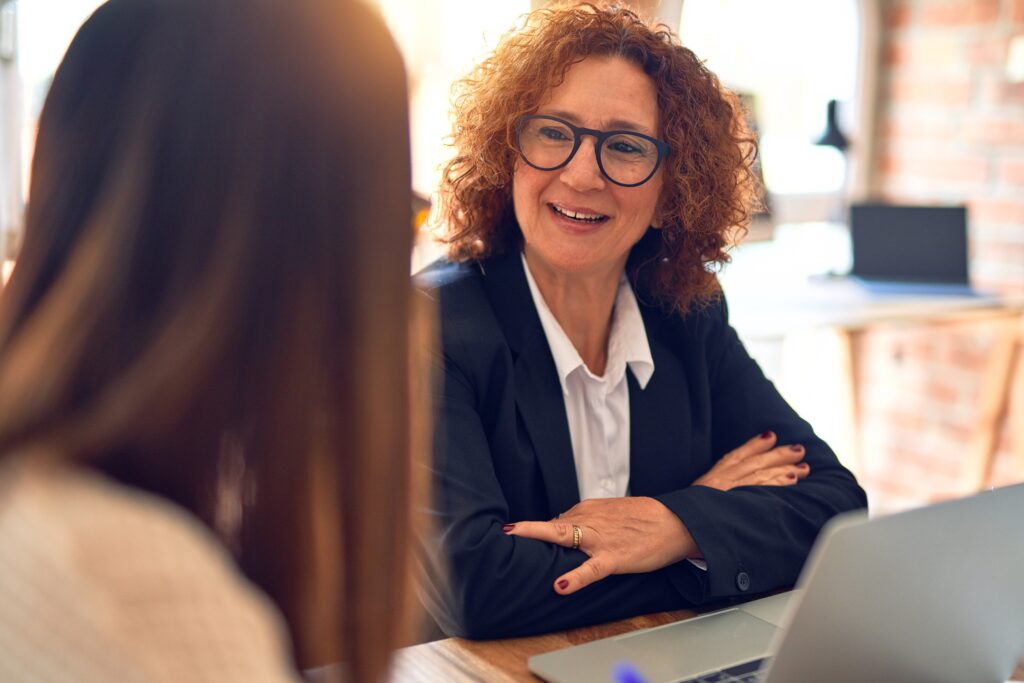 Two businesswomen smiling happy in a coaching and mentoring conversation, one more experienced worker mentoring the less experienced worker in the office.